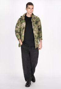 Camouflagejack met capuchon in tinten groen en beige, zwart shirt, losse zwarte jeans en zwarte laarzen. Casual, functioneel design.