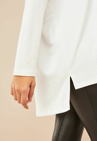Weißes Langarmoberteil mit seitlichen Schlitzen, glatter Stoffstruktur und lockerer Passform, kombiniert mit schwarzen Leggings. Fokussiert auf die Details der Ärmel und dem Saum.