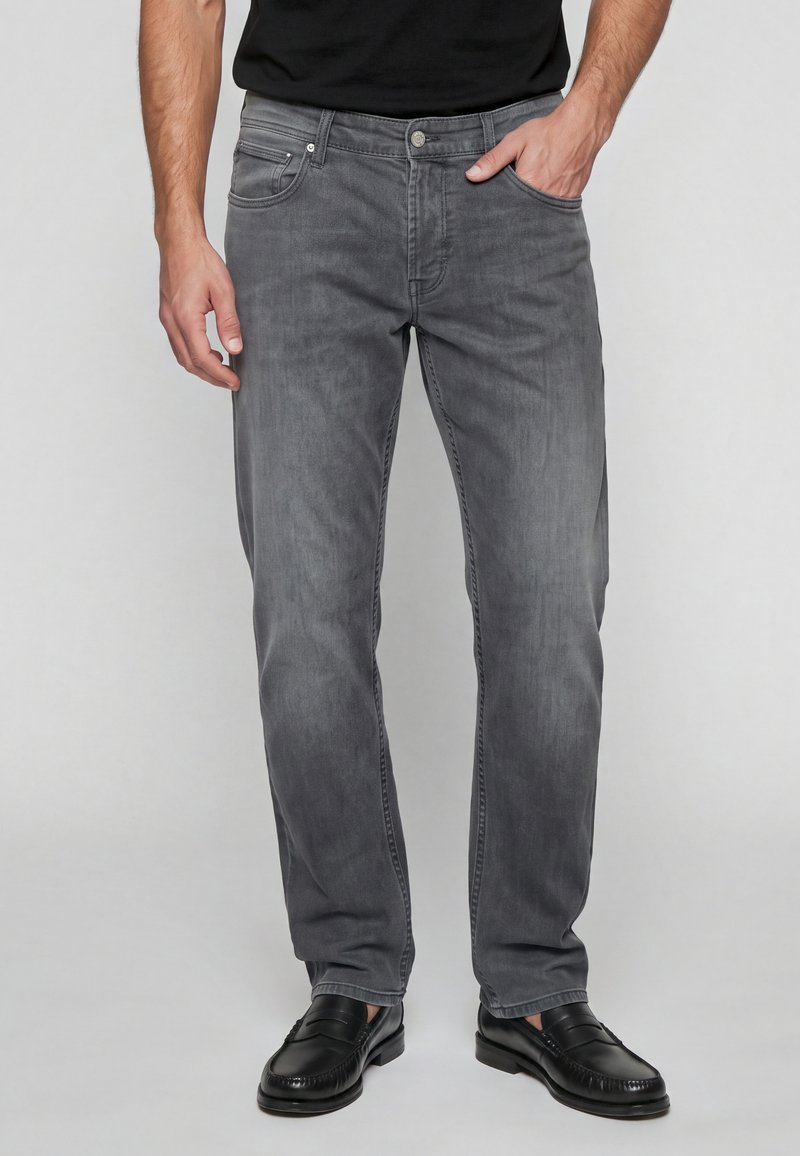 Homme portant un jean gris, debout avec une main dans la poche et des chaussures en cuir noir sur un fond clair uni.