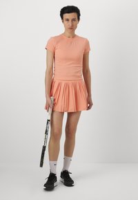 Camiseta de algodón color durazno con mangas cortas combinada con una falda plisada de durazno. Zapatillas negras y calcetines blancos de Nike, sosteniendo una raqueta de tenis.
