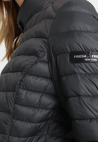 Kvinde iført en sort quiltet jakke med et "Frieda & Freddies New York" logo patch på den øverste ærme.