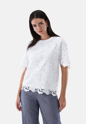 Donna con lunghi capelli scuri indossa una maglia bianca in pizzo a maniche corte e pantaloni larghi blu, in piedi davanti a uno sfondo chiaro e uniforme.