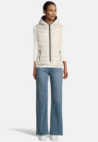 Vrouw in een mouwloze, beige, gewatteerde hooded vest over een wit geribbeld langemouwen shirt, blauwe wijde jeans en witte sneakers, staand.