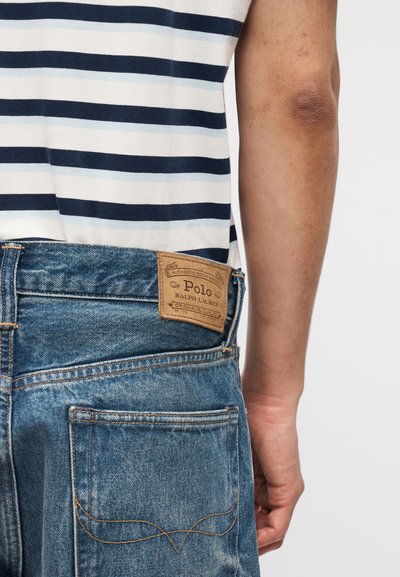 Stražnji pogled na osobu koja nosi plavu prugastu košulju uvučenu u plave traperice s kožnom oznakom Polo Ralph Lauren na struku.