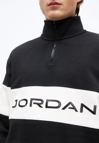 Κοντινό πλάνο ενός ατόμου που φοράει μαύρο και άσπρο φούτερ με φερμουάρ έως τη μέση και τη λέξη "JORDAN" κεντημένη στο στήθος.