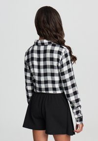 Persona con capelli lunghi e scuri, che indossa una camicia a quadri bianca e nera e pantaloni corti neri, in piedi contro uno sfondo chiaro e uniforme.