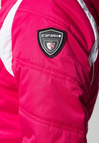 Jasnoróżowa kurtka narciarska z białymi akcentami. Posiada teksturowane wykończenie oraz czarną naszywkę z logo w kształcie tarczy z napisem "Sprzęt Narciarski" i "1996".