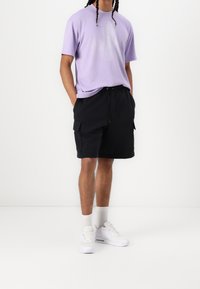 Fioletowa bawełniana koszulka z okrągłym dekoltem, zestawiona z czarnymi szortami cargo z kieszeniami. Uzupełniona białymi trampkami i skarpetkami za kostkę.