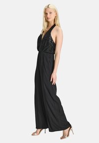 Zwarte halter-jumpsuit met een pailletten-bustier en wijde broekspijpen. Glad textuur, diep V-hals ontwerp, gecombineerd met metalen haksandalen.