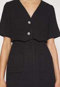Robe noire texturée à manches courtes, encolure en V profonde et deux boutons décoratifs argentés sur le haut devant.