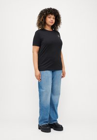 Μαύρο κοντομάνικο μπλουζάκι με μικρό λογότυπο, συνδυασμένο με απαλό μπλε τζιν σε χαλαρή εφαρμογή και μαύρα παπούτσια με χοντρή σόλα. Απλό φόντο.