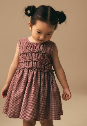 Noor tüdruk kahe kukliga, kes kannab punase-valge ruudulist varrukateta kleiti lilledetailiga, vaatab alla beeži tausta ees.