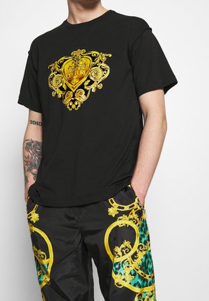 Czarny t-shirt z dużym, złotym, ozdobnym wzorem serca z misternymi detalami. Materiał jest gładki; luźny krój z krótkimi rękawami.