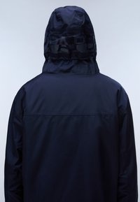 Person trägt eine dunkelmarineblaue Kapuzenjacke mit einem dezenten Markenlogo auf der Rückseite der Kapuze, von hinten vor einem einfarbigen Hintergrund gezeigt.