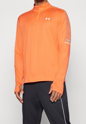 Mand iført en orange atletisk pullover med lange ærmer, kvart lynlås og tommelfingerhuller, kombineret med sorte træningsbukser med hvid stribe.