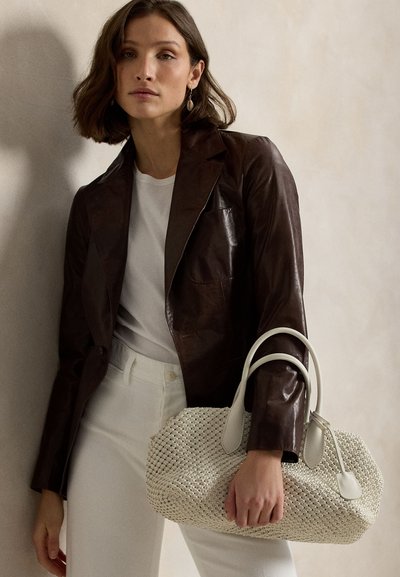 Nő sötétbarna bőrből készült blézerben és fehér nadrágban, kezében nagy, kétszeres fogantyújú, krémszínű fonott kézitáskával, semleges háttér előtt.