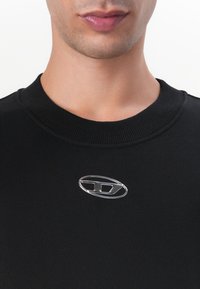 Sweatshirt noir avec un col côtelé et un logo métallique argenté au centre. Texture de tissu lisse avec un design ajusté.