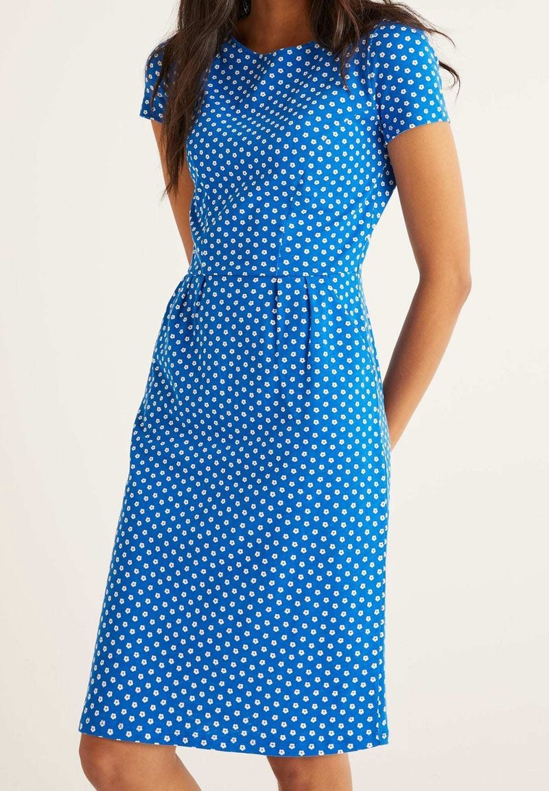 Femme portant une robe bleue mi-longue à manches courtes avec un petit motif floral blanc sur un fond clair uni.