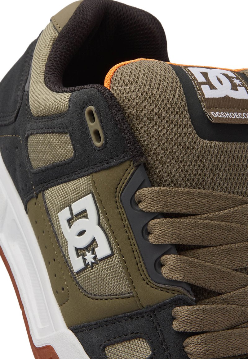 Schwarze und olivgrüne Sneaker aus strukturiertem Mesh und Wildleder, mit auffälligem Logo und orangefarbenem Akzent. Robuste Gummisohle mit Schnürsenkeln.