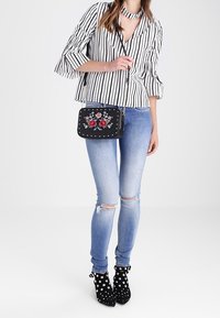 Borsa a tracolla nera con ricami floreali e borchie argentate, indossata con una blusa a righe, jeans strappati blu e stivali con tacco neri a pois.