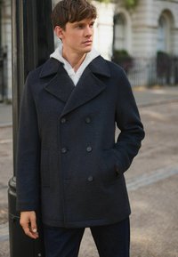 Young man wearing a dark double-breasted coat over a light hoodie, standing outdoors near a lamppost on a city street.