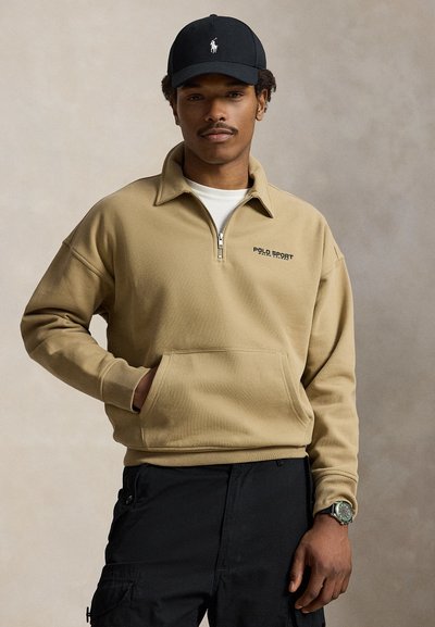 Άνδρας που φοράει μπεζ sweatshirt με φερμουάρ μέχρι το στήθος και κείμενο "Polo Sport", μαύρο καπέλο, μαύρο παντελόνι cargo και ρολόι χειρός, στέκεται μπροστά σε ουδέτερο φόντο.