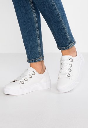 Jambes en jeans bleus portant des baskets à plateforme blanches avec de larges lacets en satin, debout sur un sol gris clair.