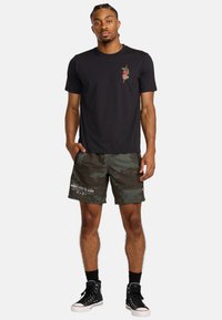 Zwarte t-shirt met een kleine kleurrijke graphic op de borst, gecombineerd met donkergroene camouflage shorts met tekst en logo, afgewerkt met zwarte sneakers.