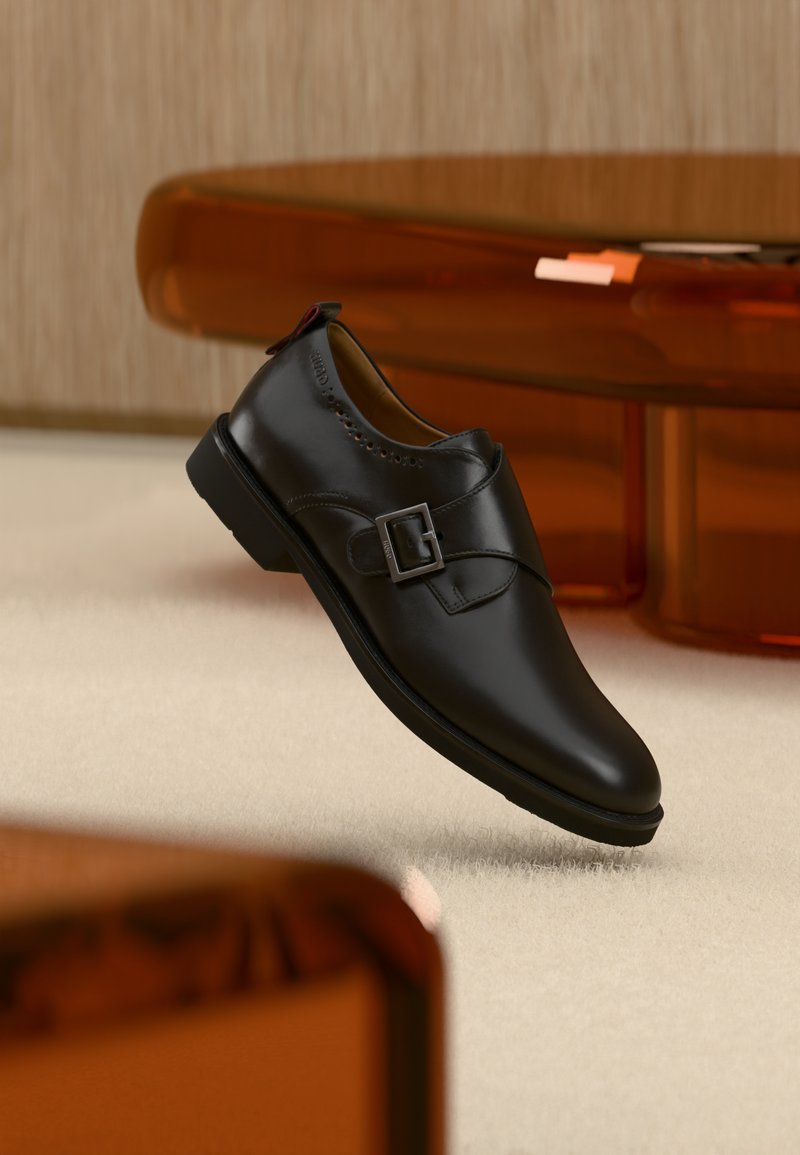 Μαύρο παπούτσι monk strap από δέρμα με ασημένια αγκράφα, αιωρούμενο πάνω από μπεζ χαλί με καφέ διαφανή έπιπλα στο φόντο.