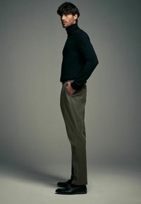 Pull en col roulé noir, pantalons vert olive et mocassins noirs avec détail en métal. Silhouette ajustée, texture lisse, design minimaliste.