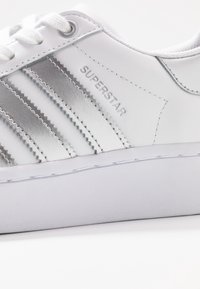 Bílé tenisky se stříbrnými metalickými pruhy, gumovou podrážkou a texturovaným svrškem. Mezi funkce patří perforované detaily a vyšívané logo.