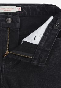 Jeans in denim nero con una vestibilità dritta, caratterizzati da una chiusura con zip dorata, bottone argento e un'etichetta bianca con la taglia 12. Dettagli del tessuto testurizzati visibili.