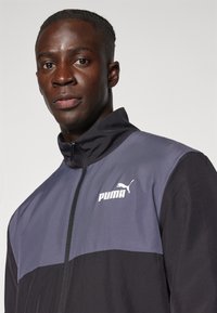PUMA-Jacke mit einem Farbblockdesign in Grau und Schwarz, hohem Kragen und Reißverschluss; Logo auf der oberen linken Seite angebracht.