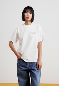 Hvid bomulds t-shirt med korte ærmer og rund halsudskæring, prydet med et lille trykt logo på venstre brystlommeområde. Båret med løse blå jeans.