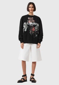 Fekete pulóver virágmintával és "ALLSAINTS LONDON" felirattal, fehér térdig érő rövidnadrággal és fekete pántos szandállal párosítva.