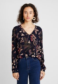 Blomstrig blus i marinblått tyg med långa ärmar, V-halsringning och färgglada växtmönster i rosa, röd och grön. I kombination med blå denimjeans.