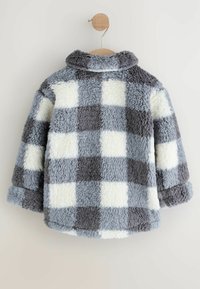 Veste en polaire à carreaux gris et blancs avec une texture douce et duveteuse, présentée suspendue à un cintre en bois sur un fond uni.