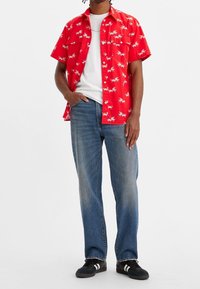 Homme portant une chemise rouge à manches courtes avec des imprimés blancs d'insectes sur un t-shirt blanc, un jean bleu et des baskets noires avec des bandes blanches.