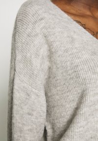 Pull en maille côtelée gris clair avec un col en V et une coupe légèrement ample. Présente une texture douce et des détails de couture visibles.