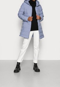 Osoba ubrana w jasnoniebieską kurtkę puchową, czarny golf, białe spodnie z mankietami i czarne masywne botki, stoi na jednolitym tle.