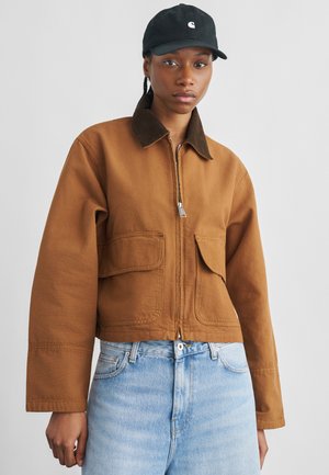 Jeune personne portant une veste marron à fermeture éclair avec de grandes poches avant, un jean bleu clair et une casquette de baseball noire sur un fond uni.