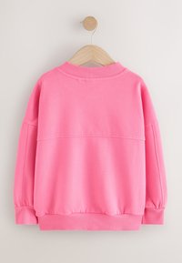 Sudadera rosa de manga larga con puños, dobladillo y cuello acanalados colgada en una percha de madera contra un fondo claro liso.