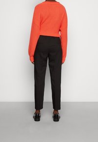 Suéter de punto naranja brillante con un corte holgado, combinado con pantalones negros a medida y zapatos negros brillantes, visto desde la parte de atrás.