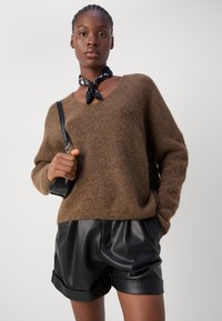 Bruin gebreid sweater met V-hals, gecombineerd met zwarte leren shorts met omgeslagen manchetten. Model houdt een kleine zwarte handtas vast.