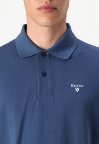 Tumesinine polo särk tekstureeritud kangast, kahe musta nupuga ja väikese valge tikandiga logoga vasakul rindkere kohal.