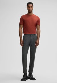 Man in een roestkleurig overhemd met korte mouwen, grijze geplooide broek en zwarte veterschoenen, staand tegen een effen achtergrond.