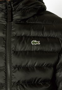 Schwarze Puffjacke mit gestepptem Design, glatter Textur, Frontreißverschluss und grünem gesticktem Krokodil-Logo.