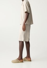 Beige linnen overhemd met korte mouwen en bijpassende knielange shorts, met subtiele verticale strepen. Bruine instap suède sandalen.