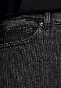 Jeans de ganga preta com um acabamento texturizado, costuras contrastantes e um pequeno bolso na frente com acessórios metálicos.