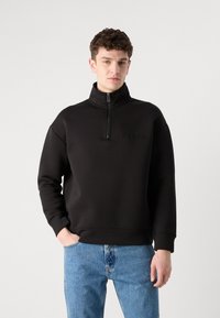 Hanorac negru cu fermoar, guler înalt, textură moale și un logo subtil pe piept, purtat cu blugi denim de culoare albastră.